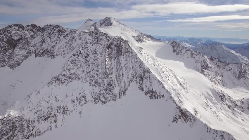 An aerial view shows vast snow-covered mountains under a cloudy sky and sunlight