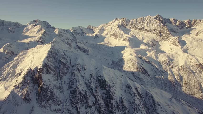 An aerial view shows snow-covered mountains under a clear sky and sunlight