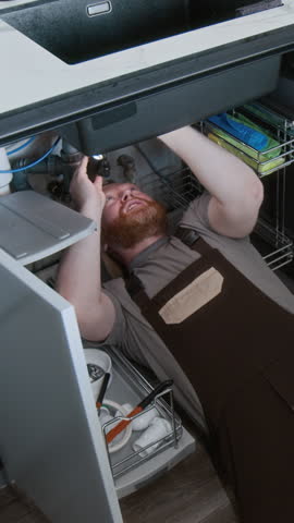 Vertical shot of man as maintenance worker or plumber inspecting kitchen sink with flashlight while lying on back and wearing coveralls