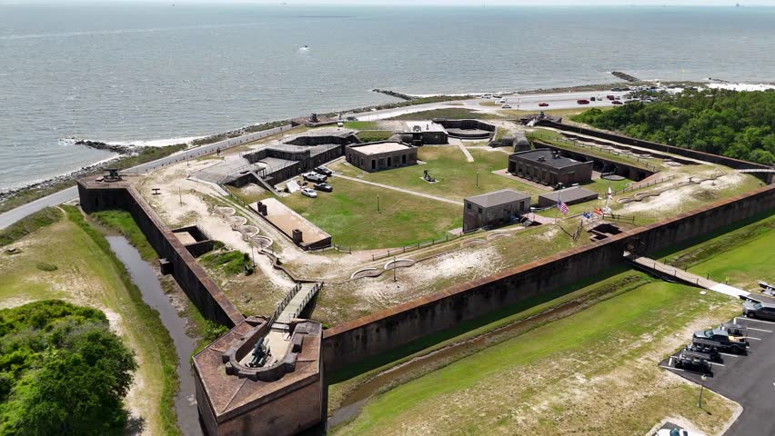 Aerial view of a historic fort surrounded by water, showcasing its large, star-shaped structure with multiple buildings and grassy areas. Nearby roads and parking areas are visible.