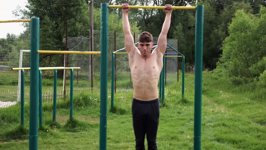 Strong guy training abdominal muscles on horizontal bar at sports ground. Young sportsman exercising on pull-up bar outdoor. Sporty athlete working out at nature. Concept of sportive lifestyle