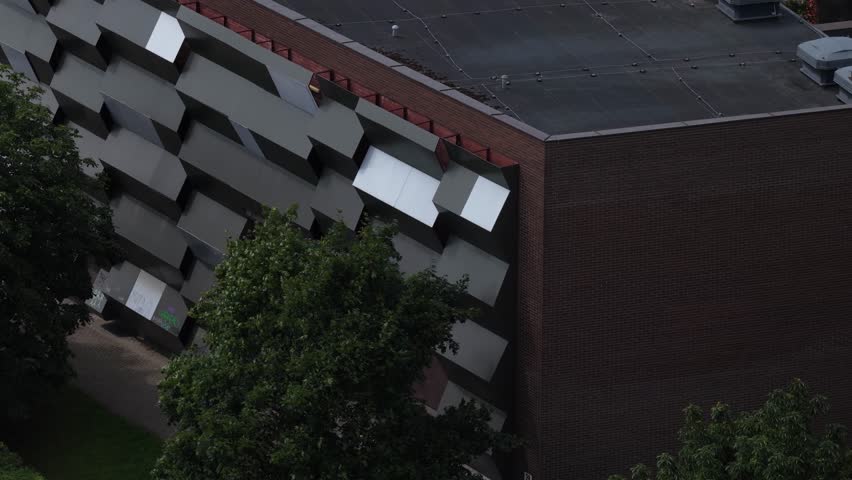 Drone shot of a building with a bold geometric facade featuring staggered cubic metal elements and a red brick top frame, surrounded by trees.