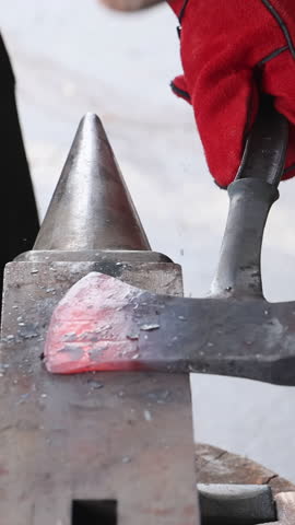An artisan shapes glowing axe making on an anvil while wearing protective gloves.