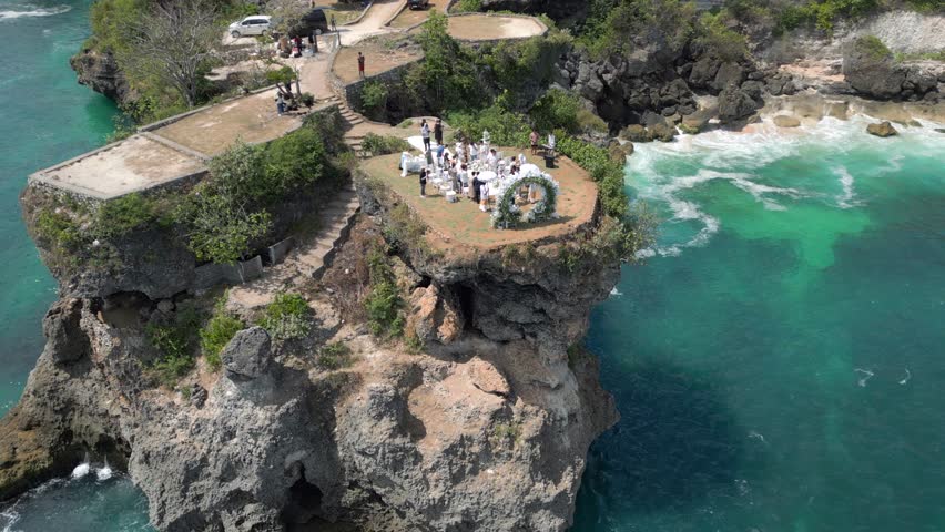 Cinematic aerial view of wedding ceremony on scenic cliff overlooking turquoise waves in Bali, Indonesia. Tropical paradise with ocean, rocks, and romantic atmosphere.