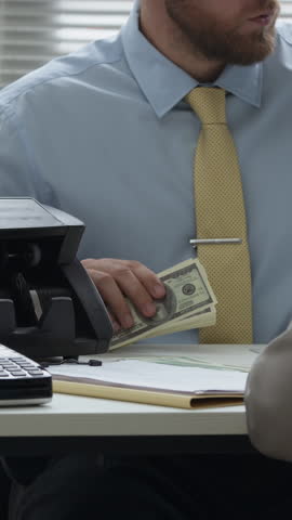 Vertical shot of male bank clerk counting paper banknotes with electronic device while communicating with client sitting at office desk