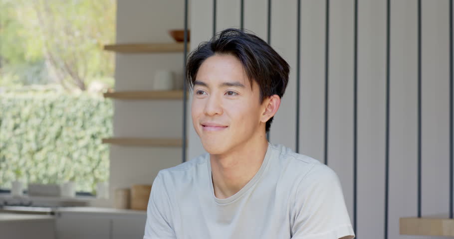 Asian man smiling, celebrating with raised fists, stretching, leaning in with grin at dining table. Joy, positivity, casual, modern, home, relaxation, wellness