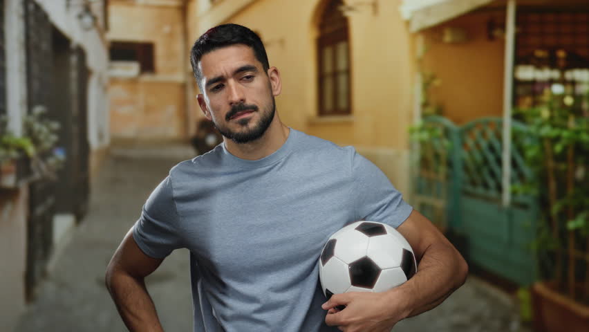 Young man with a soccer ball in hand stands confidently on an urban street with a relaxed pose, showcasing a blend of athletic style and city life against a charming town backdrop.