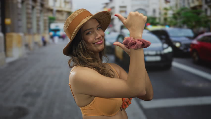 Young hispanic woman in orange bikini top and straw hat points thumb to side while smiling in a busy outdoor street; summer joy.