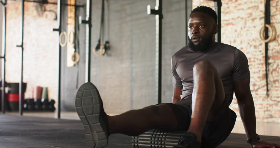 African American male placing foam roller under left calf at gym and rolling to release tension. Fitness, mobility, recovery, wellness, strength, therapy, stretch