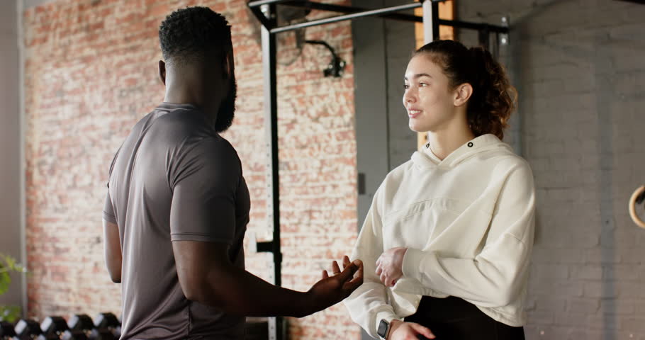 Trainer leaning forward and explaining workout movement to client at brick-walled gym beside rings. Training, mentorship, motivation, strength, fitness, wellness, teamwork