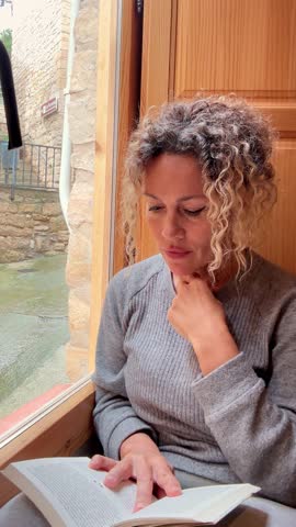 Woman at home by the window reading a book and glancing outside – Relaxed lifestyle, daylight, peaceful indoor moment, natural light, casual clothes, mature female