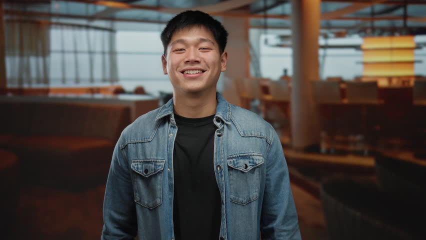 Young chinese man making glasses gesture with fingers in eyes, wearing casual denim jacket, smiling indoors at cozy coffee shop with modern decor and ambient lighting.