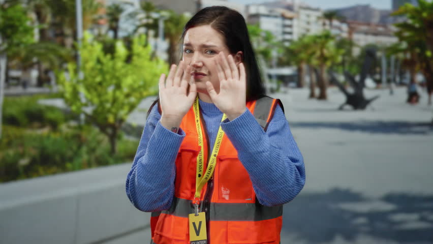Woman in orange vest covers ears from noise on busy street with green trees in the background, expressing discomfort outdoors.