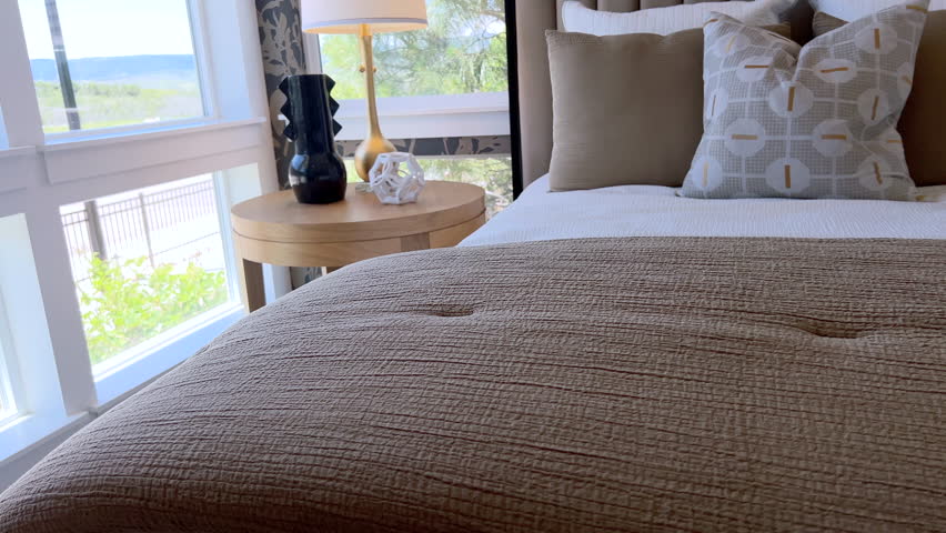 Camera frames a perfectly styled bed with decorative pillows and plush bedding. Bright natural light from large windows completes the scene.