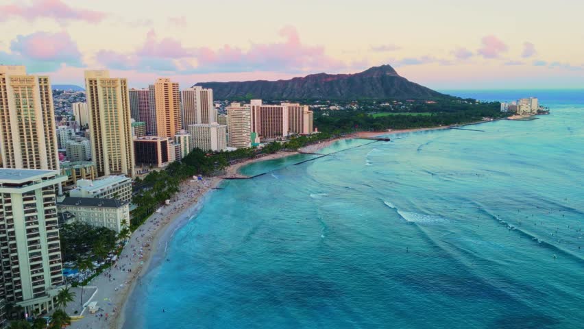 Cinematic aerial footage of Waikiki Beach at sunset, showcasing golden sand, gentle ocean waves, high-rise resorts, and palm trees, with a clear view of Diamond Head crater under a colorful sky.