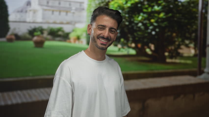 Man smiling showing teeth outdoors by a historic building with green lawn and clay pots visible behind him; happiness.