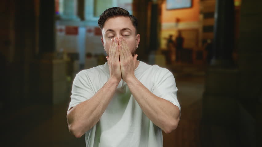 Young man with a beard stands on a city street, expressing surprise, wearing a casual white shirt in an urban outdoor environment at night, hands covering mouth.