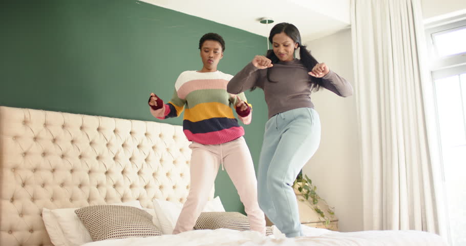 Diverse female friends stepping onto tufted bed and bouncing, swinging arms to off-screen music. Dance, friendship, entertainment, lifestyle, casual, joyful, movement