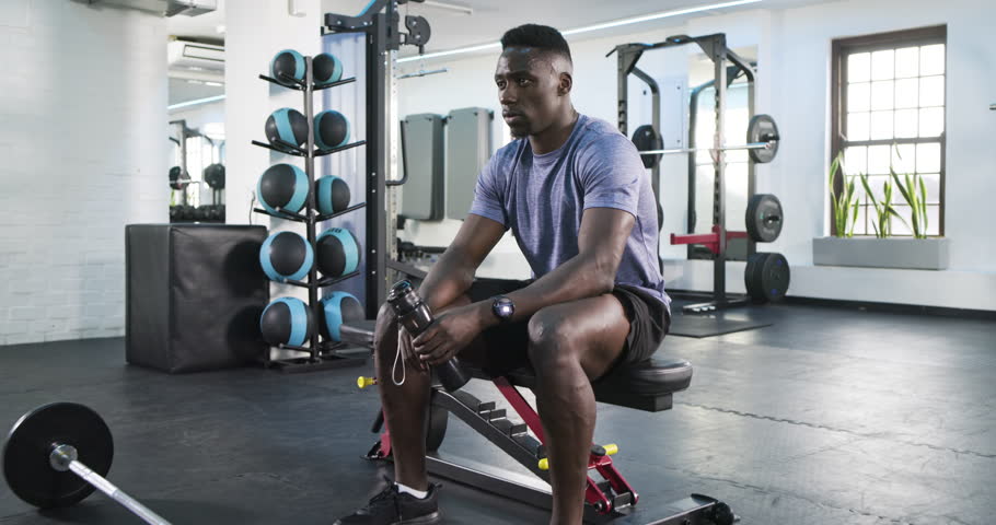 African American man sipping water from water bottle on gym bench after workout, catching breath. Athlete, fitness, hydration, strength, perseverance, determination, modern