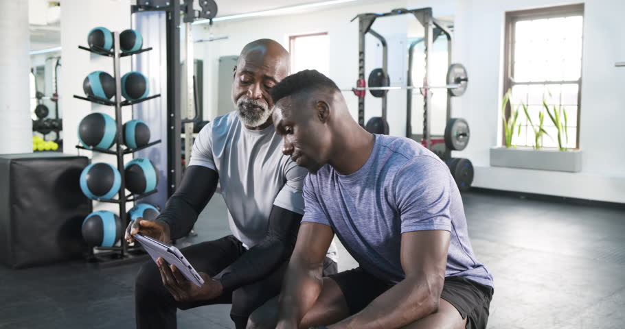 African American trainer tapping tablet, explaining routine while client watching on gym bench. Fitness, training, health, coaching, motivation, technology, strength