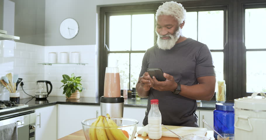 Senior African American man scrolling smoothie recipe on phone, blending in jar at kitchen island. Health, nutrition, cooking, countertop, wellness, lifestyle, domestic