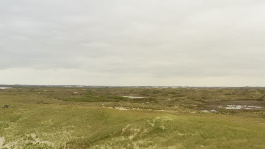 Sweeping drone shot over a massive coastal dune landscape, capturing two people biking on a path with distant water under a cloudy sky