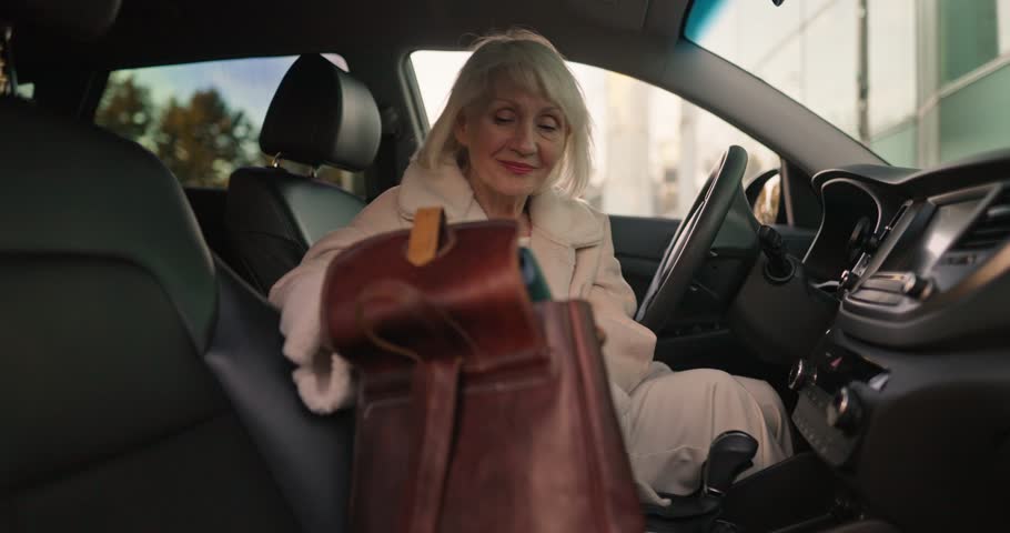 Mature woman in white coat sitting on front seat of her car and rummaging in her purse while relaxing during business trip in city. Mature wealthy woman on business trip