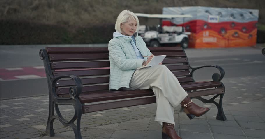Zoom in Elderly female artist with white hair marking a landscape and making sketches in her sketchbook while sitting on a park bench. Elderly woman draws Outdoor