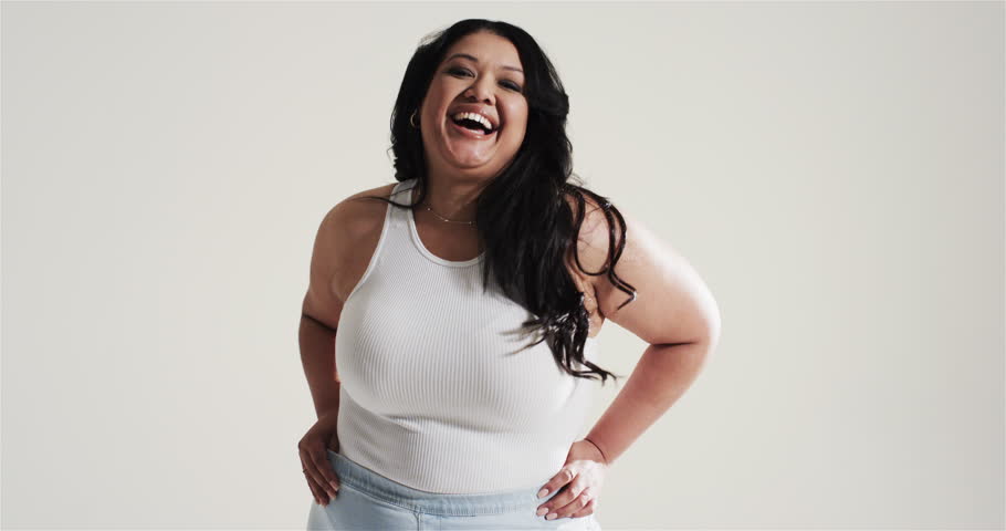 Woman wearing white tank top, jeans smiling at camera flipping hair at gray backdrop. Confidence, fashion, casual, portrait, elegance, lifestyle, minimalism