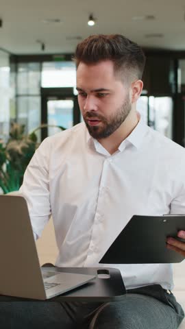 Caucasian young businessman using laptop and working with documents, analyzing information, reading papers prepare financial report. Male entrepreneur in formal analyzing graph in modern office lobby.