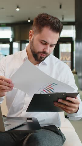 Caucasian young businessman using laptop and working with documents, analyzing information, reading papers prepare financial report. Male entrepreneur in formal analyzing graph in modern office lobby.