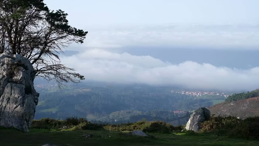 A stunning landscape of green hills and a distant town partially hidden by clouds, viewed from a high point with a large rock and a tree in the foreground. The serene scene captures the tranquility an