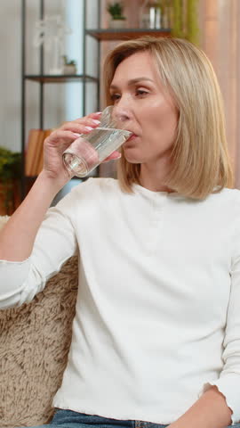 Middle-aged woman drinks filtered water on home sofa, smiling calmly, showing good lifestyle habit. Adult lady enjoys hydration, supporting body and wellness with positive mindful self-care practice.