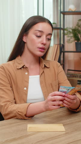 Young woman at home table counts euro income cash bills, slips part into envelope planning future purchases savings. Caucasian girl smiles quietly feeling secure about money, personal financial goals.