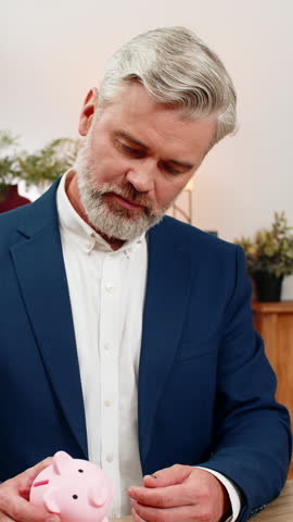 Middle-aged businessman at home office opens piggy bank empties coins realizing funds nearly gone. Freelancer guy at table counts all coins adjusts budget calculating monthly expenses seeking savings.