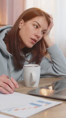 Exhausted lazy young girl freelancer working on laptop at table on sofa looking tired at the end of workday holding head with one hand. Documents, cup of coffee are on table. Woman working from home