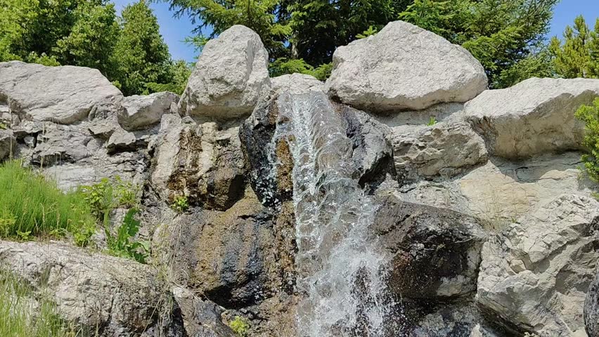 The video shows a waterfall or a rapid stream of water cascading down a rocky slope. The water moves quickly, creating white streams and sprays that contrast with the dark wet rocks. In the background