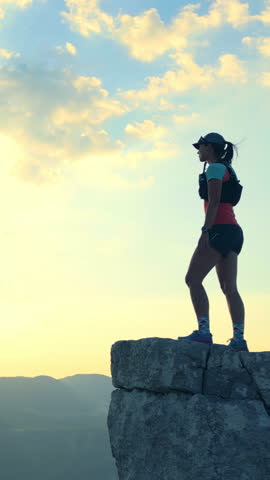Determined female runner triumphantly stands on a rugged cliff, gazing into the horizon. Dressed in athletic gear, she embodies cross-country running spirit amidst a serene, expansive landscape.