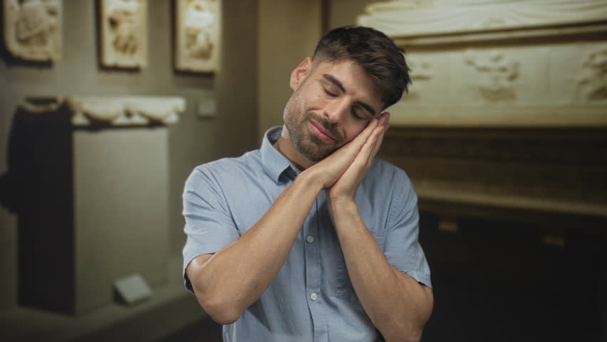 Young hispanic man rests head on hands in museum gallery lined with classical antiquities and marble reliefs; serenity.