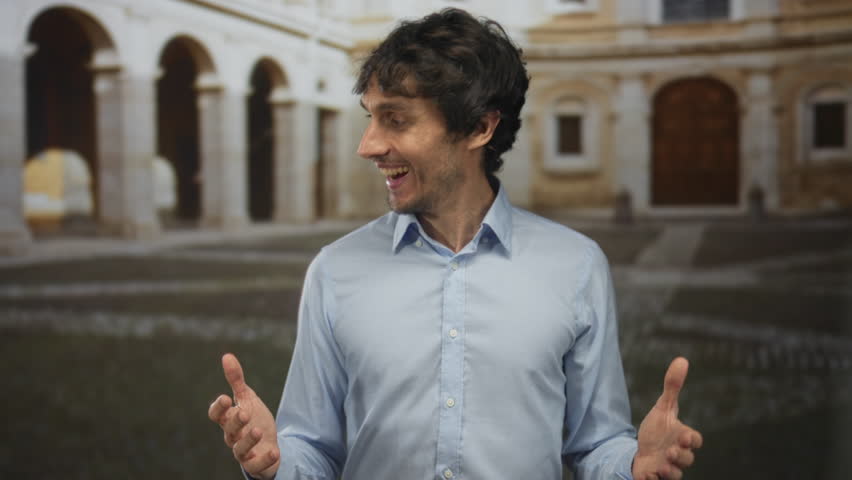 Man spreads open hands and opens mouth wearing a casual blue shirt under bright sunlight before building; enthusiasm.