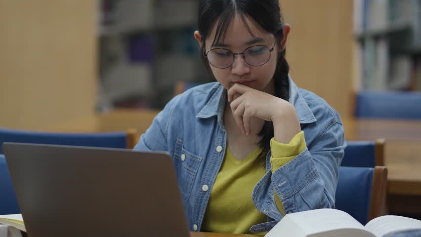 Young Asian woman, early 20s, wearing glasses and casual clothes, studying in a library using a laptop and book. Ideal for concepts like education, learning, focus, university, or academic life.