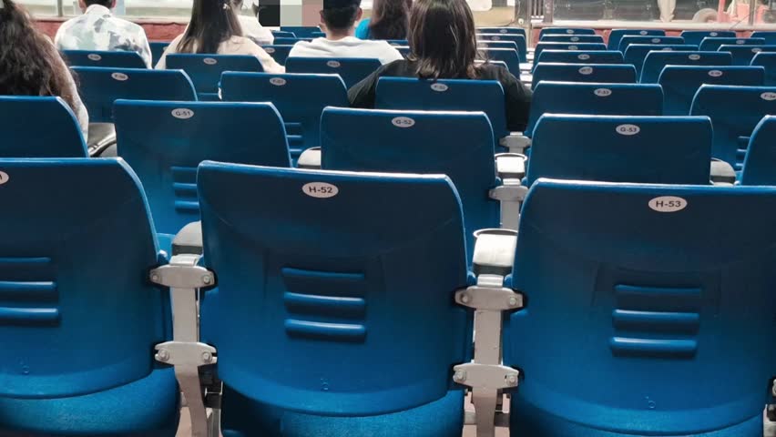 Backview video of spectators sitting on blue stadium chairs, watching a live sports event. The scene captures audience anticipation and collective focus from multiple angles