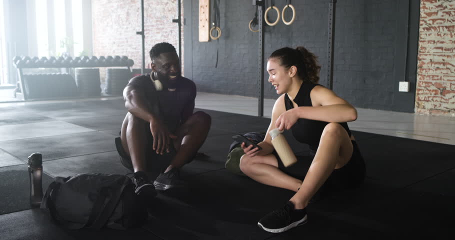 Diverse friends breathing post workout at gym, scrolling phone reviewing stats and sipping shake. Fitness, camaraderie, activewear, wellness, outdoor, recovery, casual