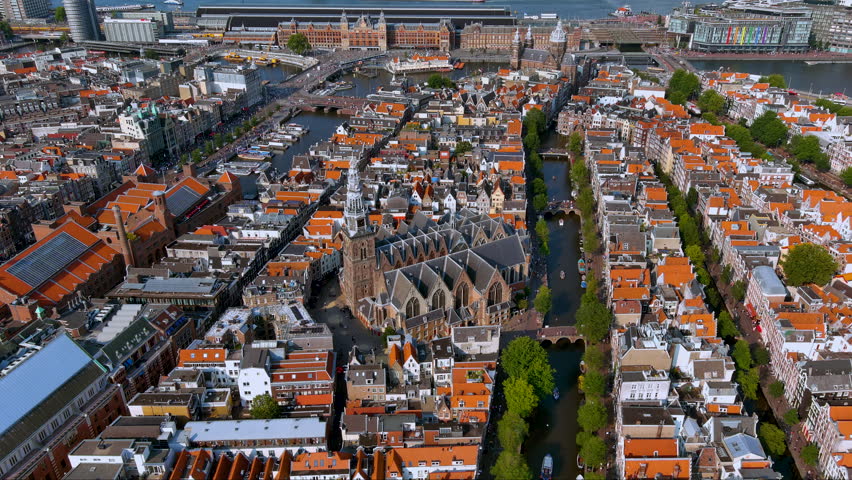 4K drone view of Amsterdam’s historic core : the Gothic Oude Kerk ringed by canals and gabled houses, with Amsterdam Central Station and the IJ waterfront in the distance.