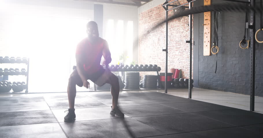 African American male gripping rope at gym squatting swinging it then crouching catching breath. Strength, conditioning, workout, fitness, gym, training, athletic