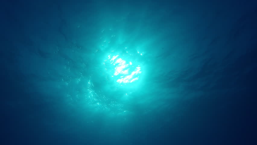 Bottom-up view on sun from blue abyss, light filters down through blue water surface. Sunbeams from deep ocean. Shining sunlight under water, with waves on top waters. Shimmering sunshine undersea