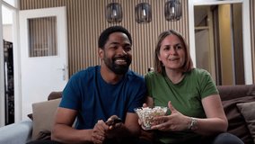 Happy interracial couple watching TV while eating popcorn at home, enjoying quality time together in warm living room atmosphere, 4k video footage. - Powered by Shutterstock - Get 15% off with code: PIKWIZARD15