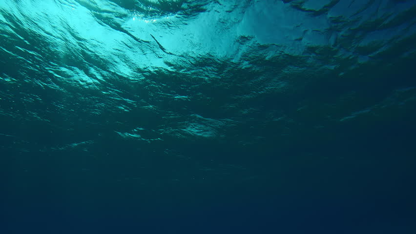 Bottom view on sea waves on the surface of water with underwater sunlight shines in the deep, View on sunlight from blue abyss, Slow motion of storm waves with sunbeams in the blue depth of Ocean