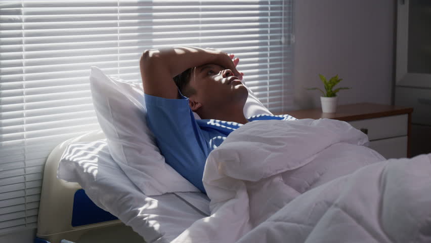 Patient experiencing severe headache lying motionless in hospital bed, sunlight filtering through partially closed window blinds, expressing physical and emotional distress