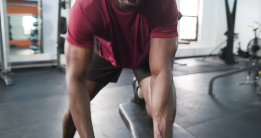 Approaching bench, African American man performing bent-over dumbbell rows at gym for back strength. Fitness, strength, training, gym, workout, athletic, wellness